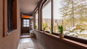 Un couloir avec une porte en bois, une petite table et des plantes en pot près de grandes fenêtres donnant sur un paysage enneigé.