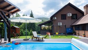 Une piscine avec une terrasse en bois, un espace de jeux pour enfants et une maison équipée de climatiseurs.
