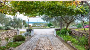 Un camí pavimentat que condueix a una entrada tancada per una reixa, envoltat de vegetació i una paret de pedra.