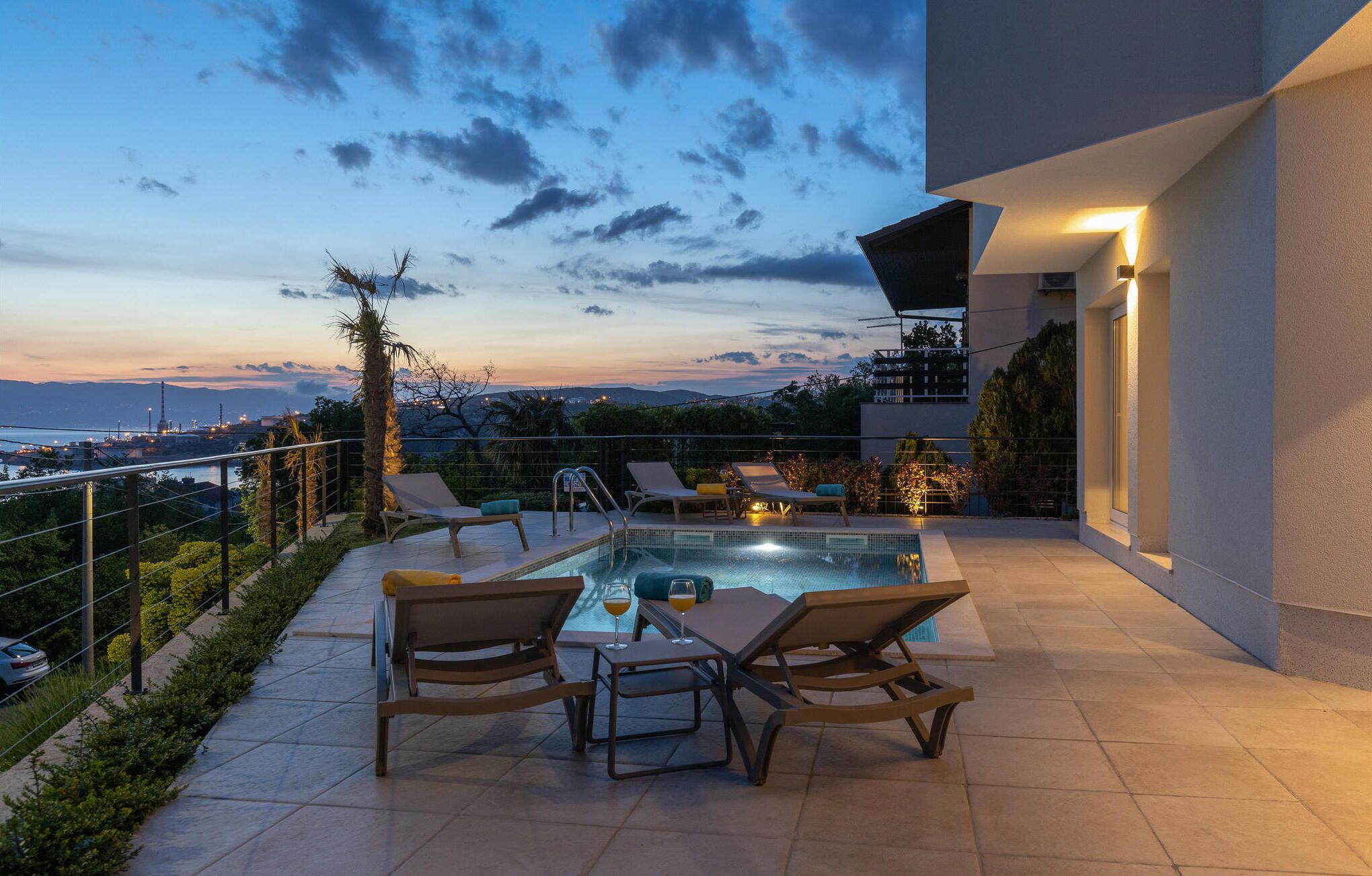 Uma área moderna de piscina ao ar livre com espreguiçadeiras e uma mesa, com vista para uma paisagem cênica de uma cidade e montanhas durante o pôr do sol.
