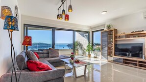 Uma sala de estar moderna com sofá, mesa de centro e TV, janelas amplas com vista para o mar e um móvel de entretenimento de madeira.