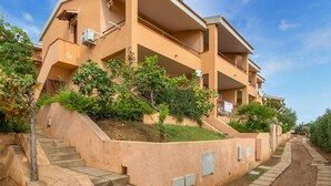 Um edifício residencial de vários andares com varandas, uma escadaria que leva à entrada e um jardim com árvores e arbustos.