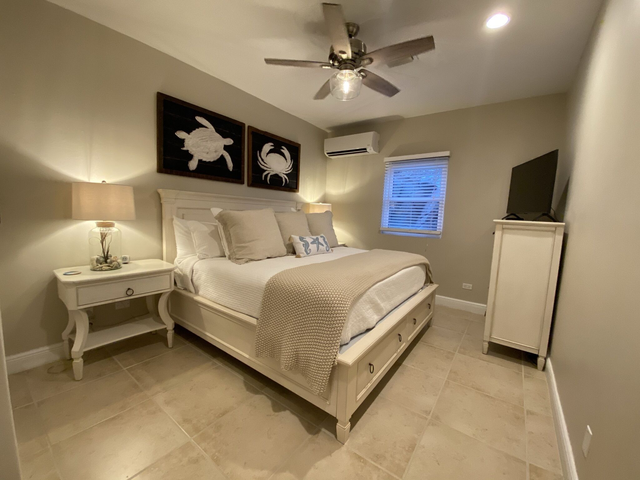 A bedroom with a bed, nightstands, a ceiling fan, an air conditioning unit, a window with blinds, and a cabinet.