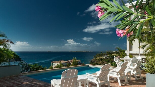 Un espace aménagé au bord de la piscine, avec des chaises et des tables blanches, offrant une vue sur une mer d’un bleu limpide et un paysage vallonné.