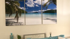 Une pièce décorée d’un grand tableau représentant une plage tropicale, d’une citation encadrée et d’un canapé turquoise agrémenté de coussins décoratifs.