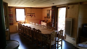 A dining area with a long wooden table and chairs, a window with curtains, and a wooden wall with a rooster painting.