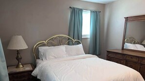 A neatly made bed with white linens, a bedside lamp, a wooden nightstand, and a dresser with a mirror. - Apt 3 Comfortable and Convenient (Masury)