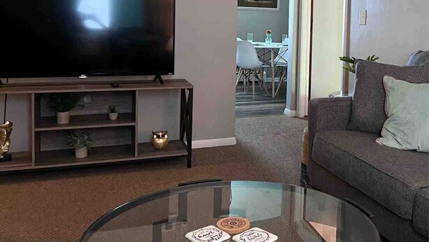 A living room with a flat-screen TV on a stand, a glass coffee table with coasters, a grey sofa, and a dining area in the background. - Apt 3 Comfortable and Convenient (Masury)