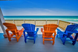 A wooden deck with four Adirondack chairs facing a beach.