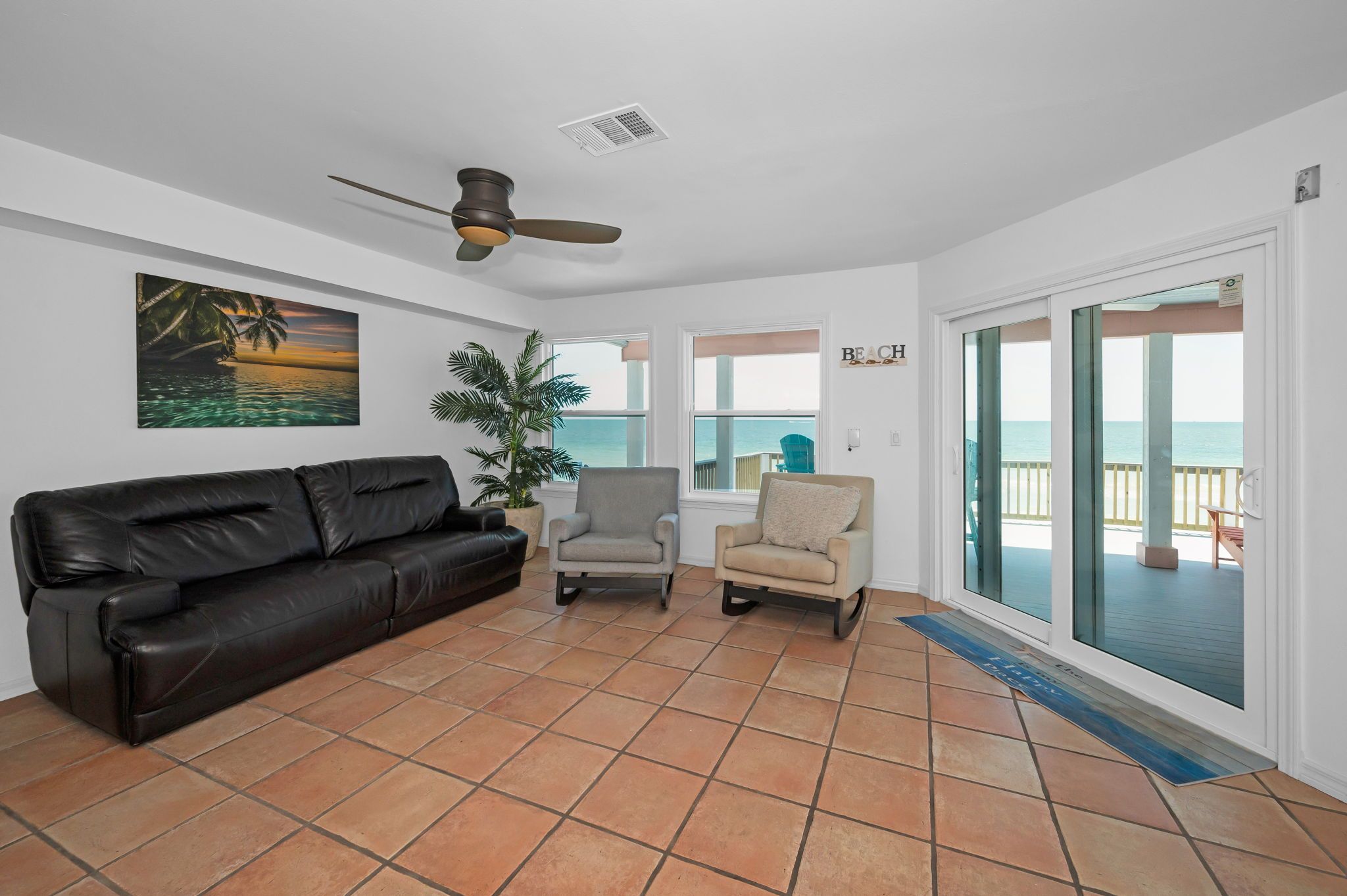 A living room with a black leather sofa, two armchairs, a ceiling fan, and a large window with a view of the beach.