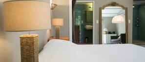 Ein Hotelzimmer mit Bett, Lampe, Korbsessel, Spiegel und einem durch eine offene Tür einsehbaren Badezimmer.