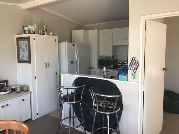 A compact kitchen with white cabinets, a refrigerator, a microwave, and a small dining area with two stools.