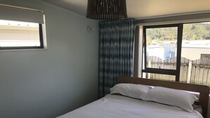 Uma cama de solteiro com roupa de cama branca em um quarto com janela, luminária na parede e cortina.
