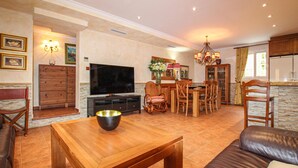 A living room with a TV, wooden furniture, a dining area with chairs, a chandelier, and a kitchen area with cabinets. - Beautiful home in Orihuela (Orihuela)