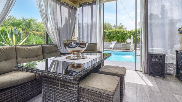 Une terrasse ensoleillée avec une table à plateau de verre, des meubles en osier et une vue sur une piscine.