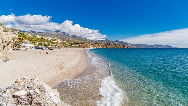 Una platja amb aigua turquesa cristal·lina, un litoral rocos i un paisatge de muntanyes i una vila de fons.