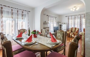 A dining area with a round table set for four, surrounded by chairs. There is a sofa and a television in the background.