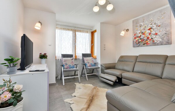Un salon comprenant un canapé, un téléviseur, un bureau avec un ordinateur et une chaise. Il y a une fenêtre avec des rideaux, un tableau au mur et un tapis sur le sol.