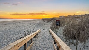Een houten looppad dat naar een strand leidt, met een zonsondergang op de achtergrond.