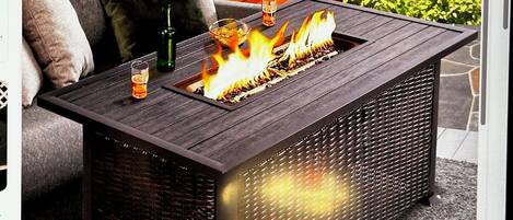 Un área de estar al aire libre rectangular con una chimenea integrada, una mesa de vidrio y un sofá acolchado con cojines.