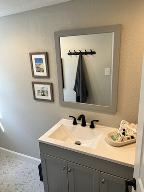 Une salle de bain avec un lavabo blanc, des robinets foncés et un miroir au-dessus. Deux tableaux encadrés sont accrochés au mur et une serviette est suspendue à un crochet.
