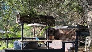 A wooden deck with a covered barbecue area, surrounded by trees. - Casita in the Oaks (Canyon Lake)