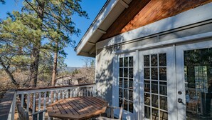 A wooden deck with a table and chairs, a white door with glass panels, and a view of trees and a distant building. - Lunar Lodge -  Forest Views | Fireplace | Smart TV (Big Bear Lake)
