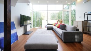 Sala de estar moderna com um sofá cinzento, uma televisão de tela plana e uma grande janela com vista para o verde lá fora.