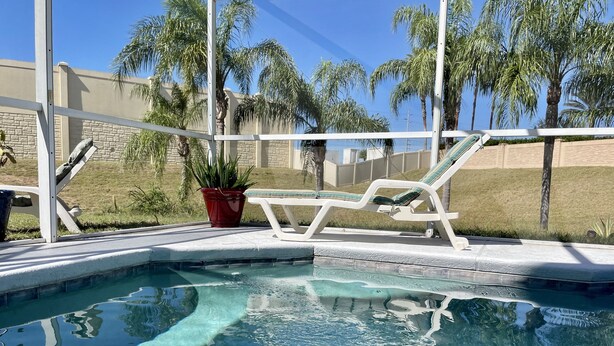 A clear pool with a white poolside area, a red potted plant, and lounge chairs.