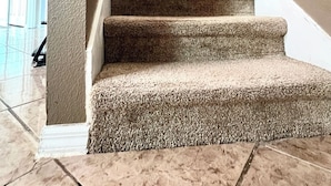 A carpeted staircase with a handrail, leading to a landing with a light switch and a white wall-mounted shelf.