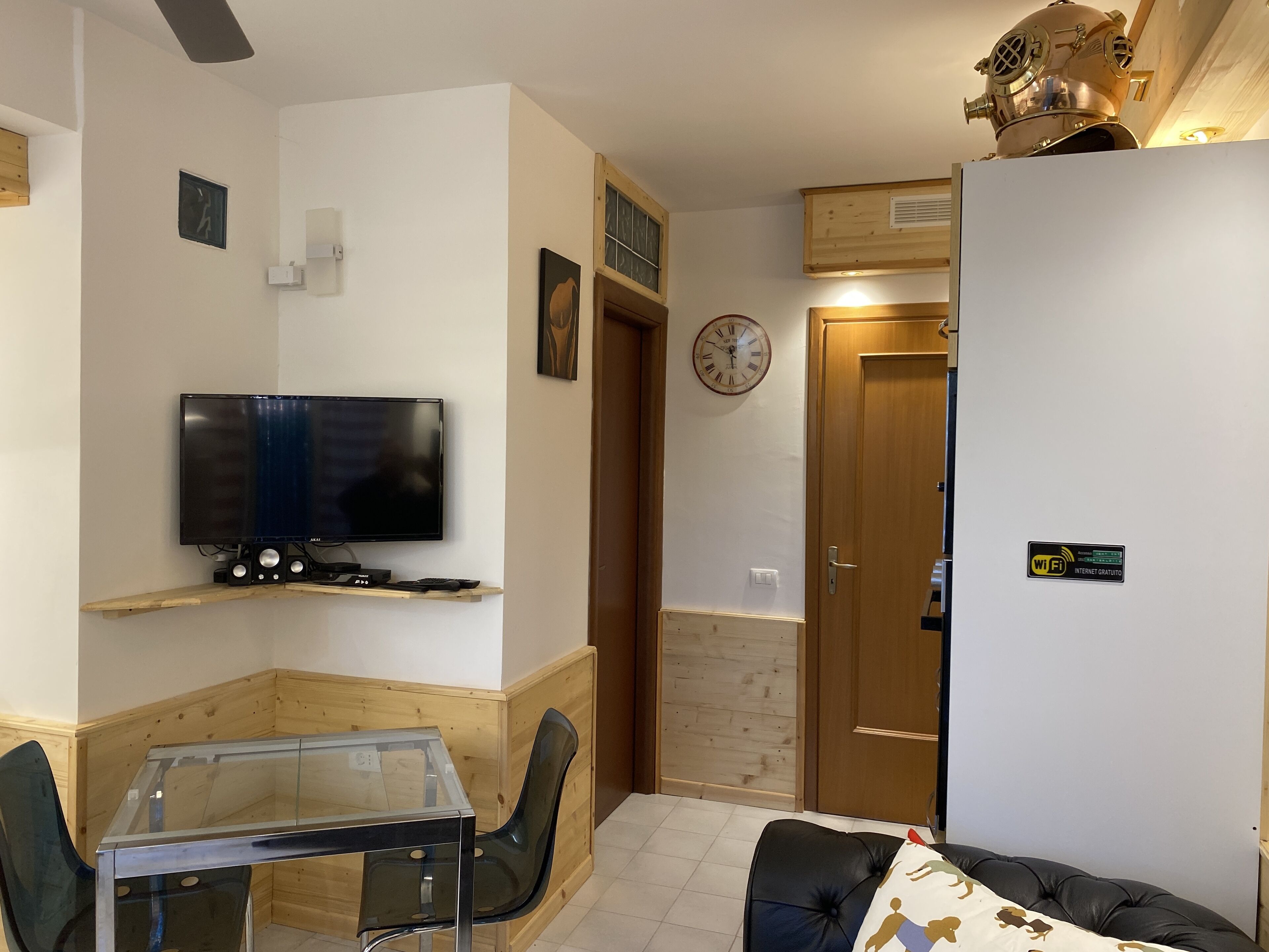 A compact living space with a TV on a wooden shelf, a glass table, a black sofa, and a wooden door with a clock.