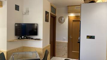 A compact living space with a TV on a wooden shelf, a glass table, a black sofa, and a wooden door with a clock.
