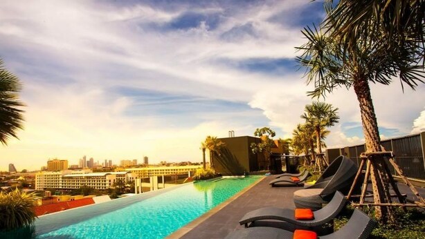 A rooftop pool with lounge chairs and palm trees, overlooking a city skyline. - The Chezz Condominium by Patsamon (Pattaya)