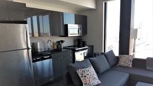 A modern kitchen with stainless steel appliances, a grey sofa with patterned cushions, and a view of the city through large windows. - Cozy Skyline Entire Condo in Downtown - Across from Bell MTS Centre (Winnipeg)