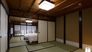 Une pièce de style japonais traditionnel, avec un sol recouvert de tatamis, un lit avec une literie blanche, des portes coulissantes et un luminaire au plafond.