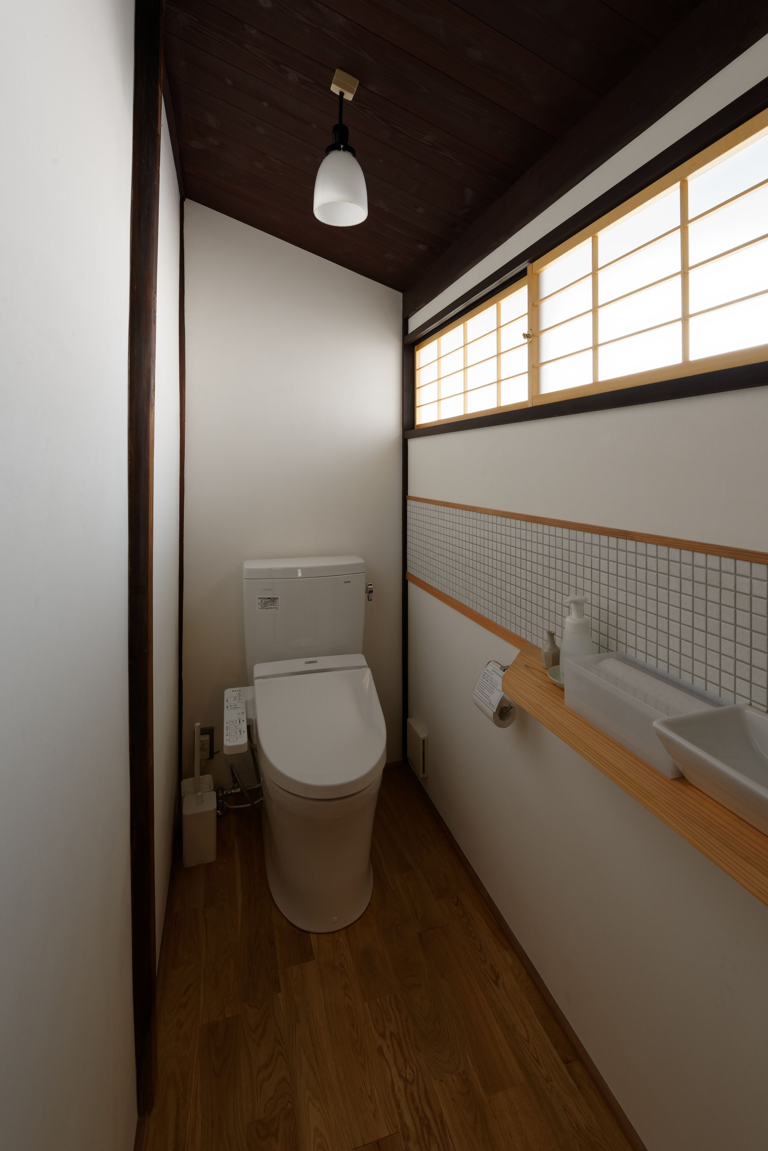 Une petite salle de bain comprenant des toilettes, une étagère en bois avec des articles de toilette, un pare-splash carrelé et une fenêtre à motifs géométriques.