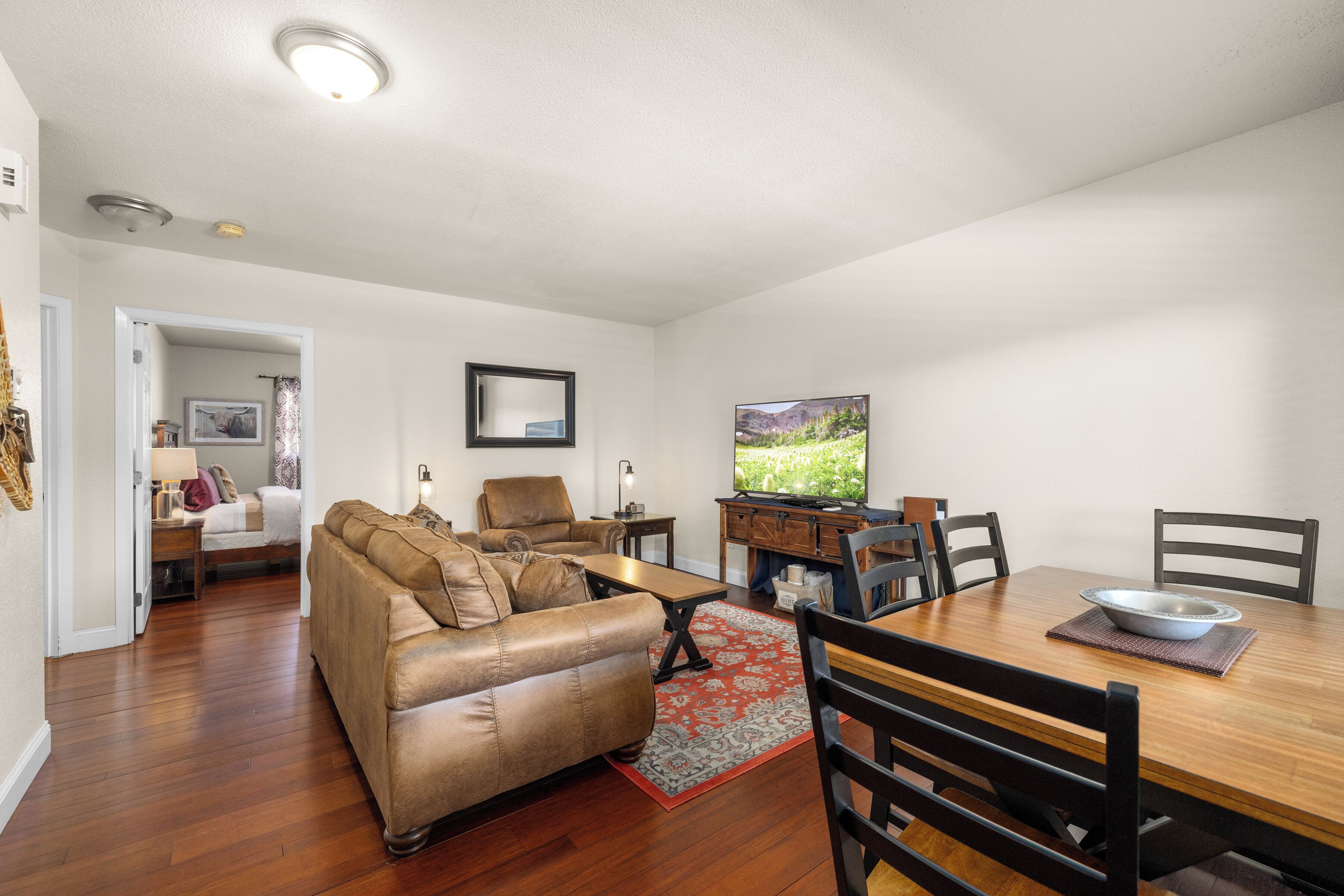 A living room with a sofa, coffee table, and dining table with chairs. There is a painting on the wall and a window with a view of greenery.