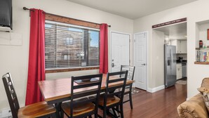A dining area with a wooden table and chairs, a window with blinds, a door, and a kitchen visible in the background. - 2 bedroom condo in Downtown Whitefish, Close To Glacier Park, Skiing And Golf (Whitefish)
