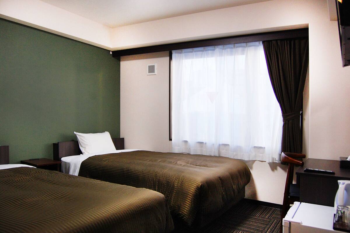 Habitación de hotel con dos camas, un escritorio, una silla, una ventana con cortinas y una mesita con lámpara.