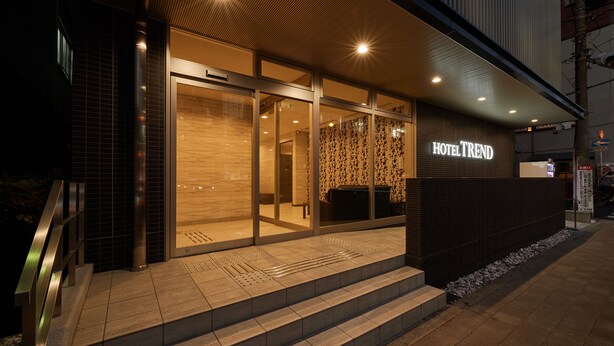 Entrada moderna de hotel com fachada de vidro, área de recepção visível e escadaria que leva à entrada.