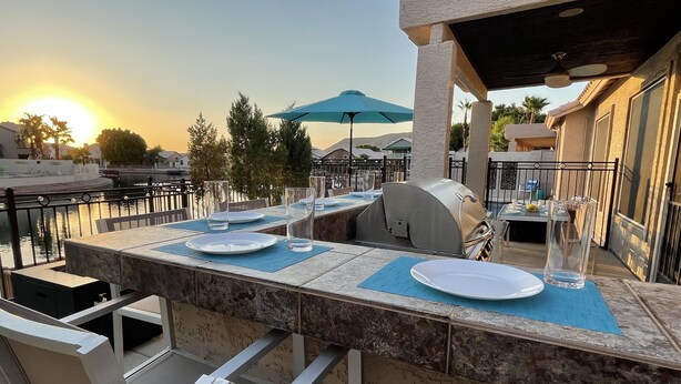 An outdoor dining area with a grill, tables, and chairs, overlooking a body of water. - Luxury Lakeside Home! Private Pool, Dock, Outdoor Kitchen, & Private Courtyard! (Glendale)
