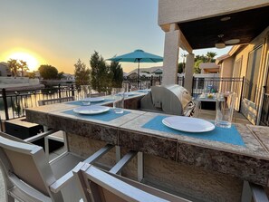 An outdoor dining area with a grill, tables, and chairs, overlooking a body of water. - Luxury Lakeside Home! Private Pool, Dock, Outdoor Kitchen, & Private Courtyard! (Glendale)