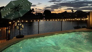 A swimming pool with a view of a sunset over a body of water, a fence with string lights, and a large umbrella. - Luxury Lakeside Home! Private Pool, Dock, Outdoor Kitchen, & Private Courtyard! (Glendale)