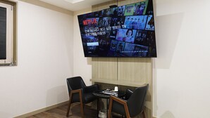 A room with a large TV mounted on the wall displaying various content. There are two black chairs and a small table with a white cup and some papers. - Mind Hotel (Seoul)