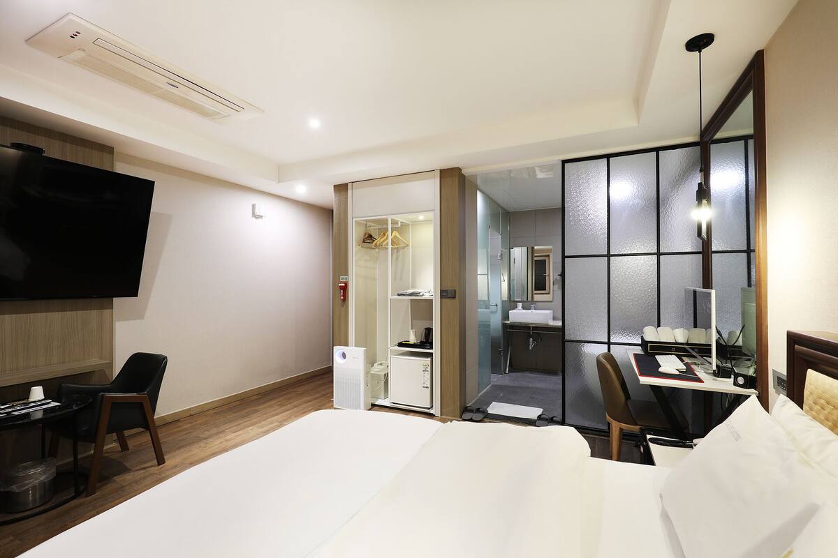 A hotel room with a bed, a desk with a computer, a TV, a wardrobe, and a bathroom visible through a glass door.