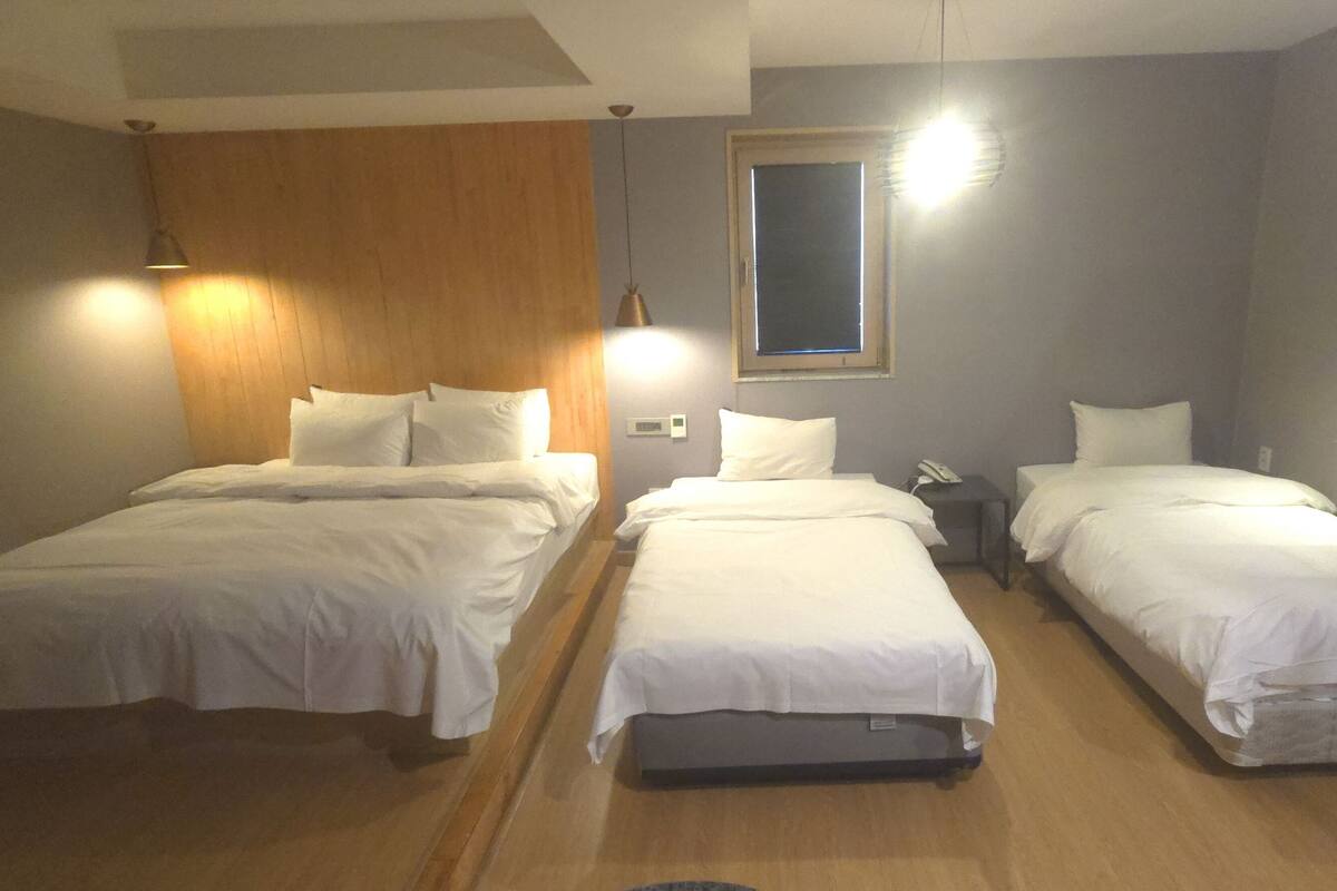 A hotel room with three single beds, wooden flooring, a wooden headboard, a small desk, and a nightstand with a lamp.
