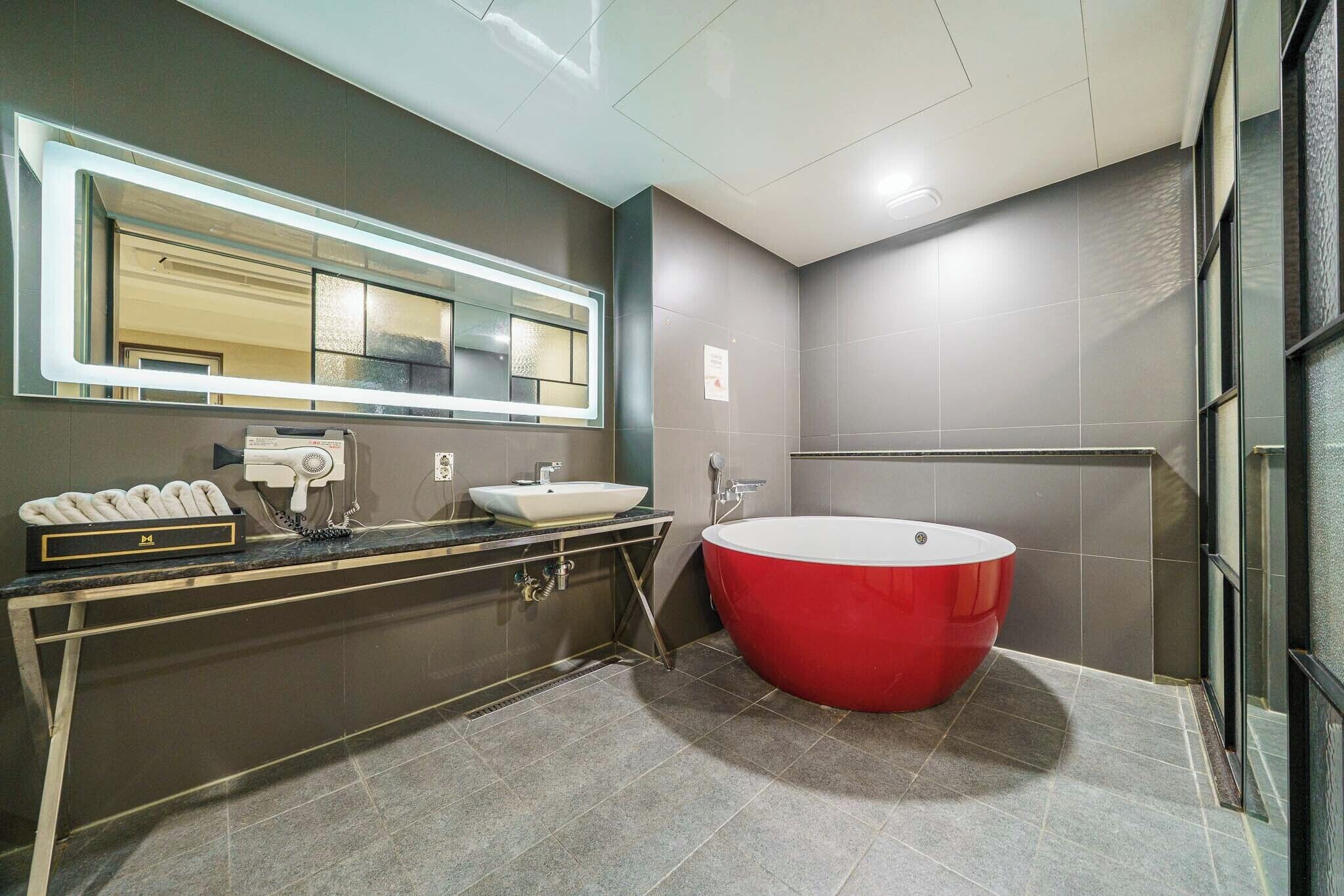 A modern bathroom with a large mirror, a red bathtub, a white sink, and a hairdryer on the vanity.