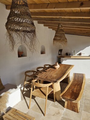 Uma área de jantar rústica em madeira, com uma mesa grande e bancos, um lustre pendurado e uma pequena cozinha com pia e bancada.
