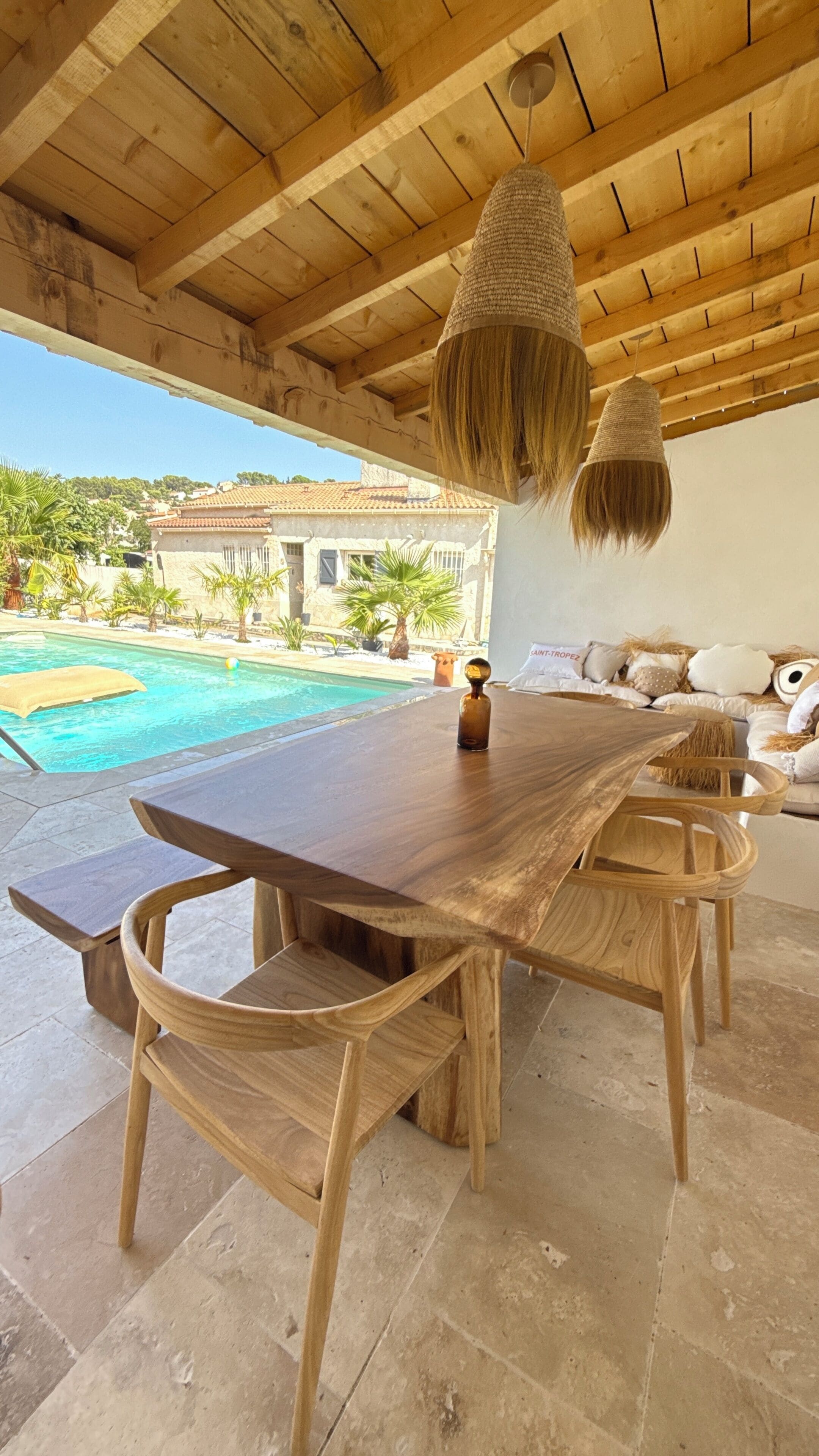 A wooden dining table with chairs, a hanging light fixture, and a view of a swimming pool and buildings in the background.