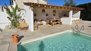 Un área de piscina con una piscina azul cristalina, una terraza de madera y un edificio blanco con techo de tejas.